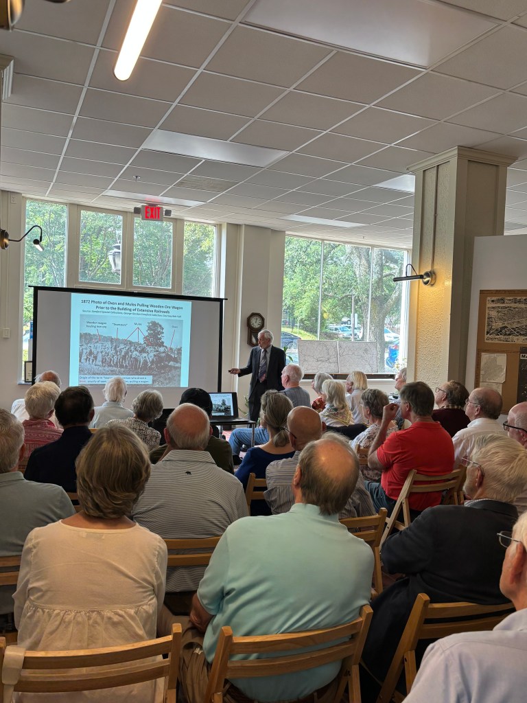 A speaker presenting to an audience in a well-lit room, with a projected historical image on the screen about railroad construction from 1872. The audience, consisting of mostly older adults, is seated and focused on the presentation.