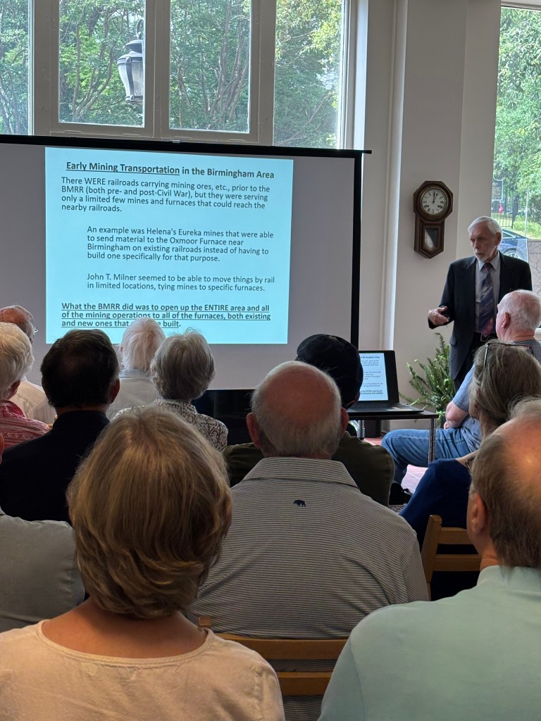 A speaker presenting information on early mining transportation in Birmingham, with a projector screen displaying text and an audience attentively listening.