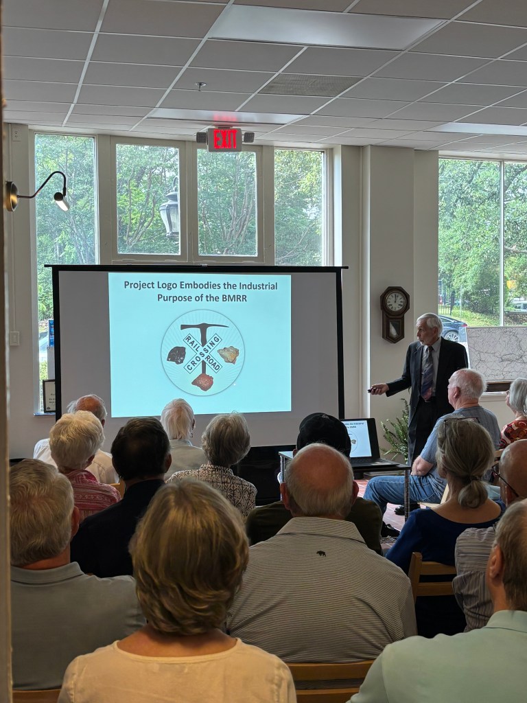 A speaker presents to an audience, displaying a slide titled 'Project Logo Embodies the Industrial Purpose of the BMRR' with visuals representing a logo related to rail and industrial themes.