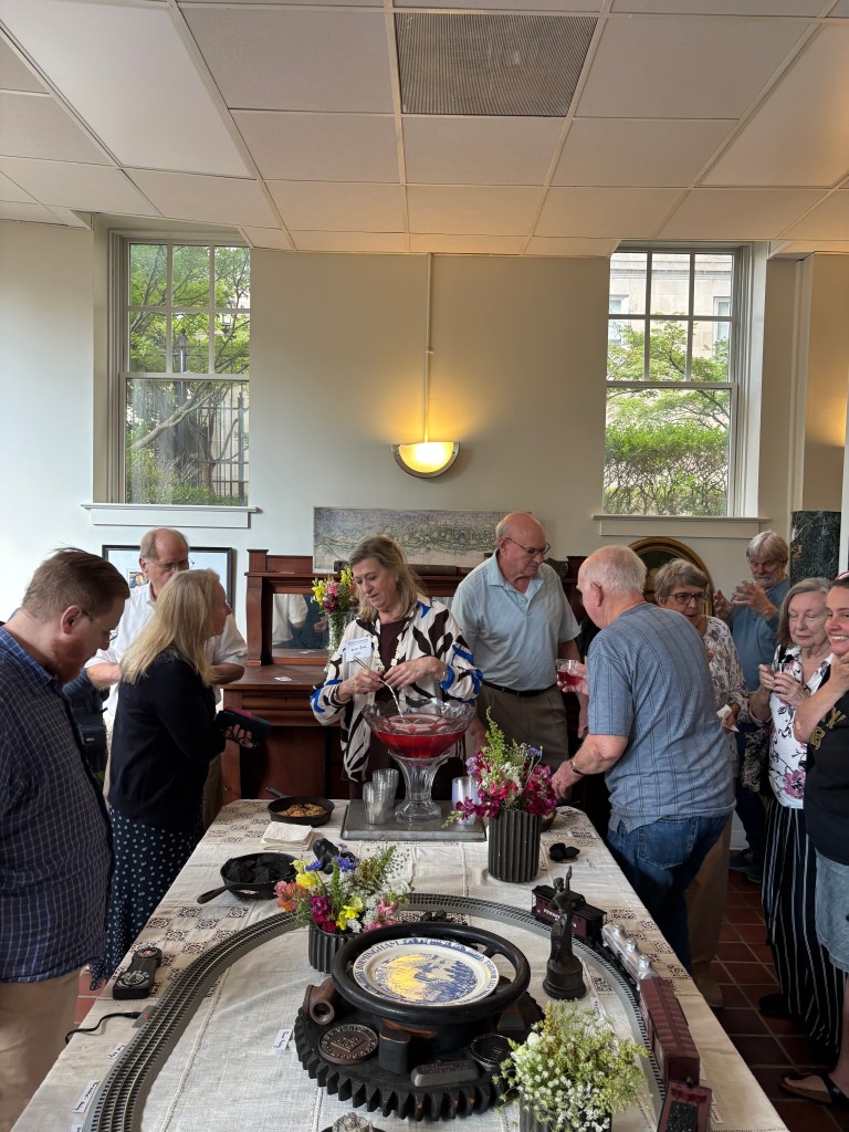 A lively gathering of people socializing at an indoor event, with a table featuring drinks, snacks, and floral decorations. A model train track is also visible in the foreground.