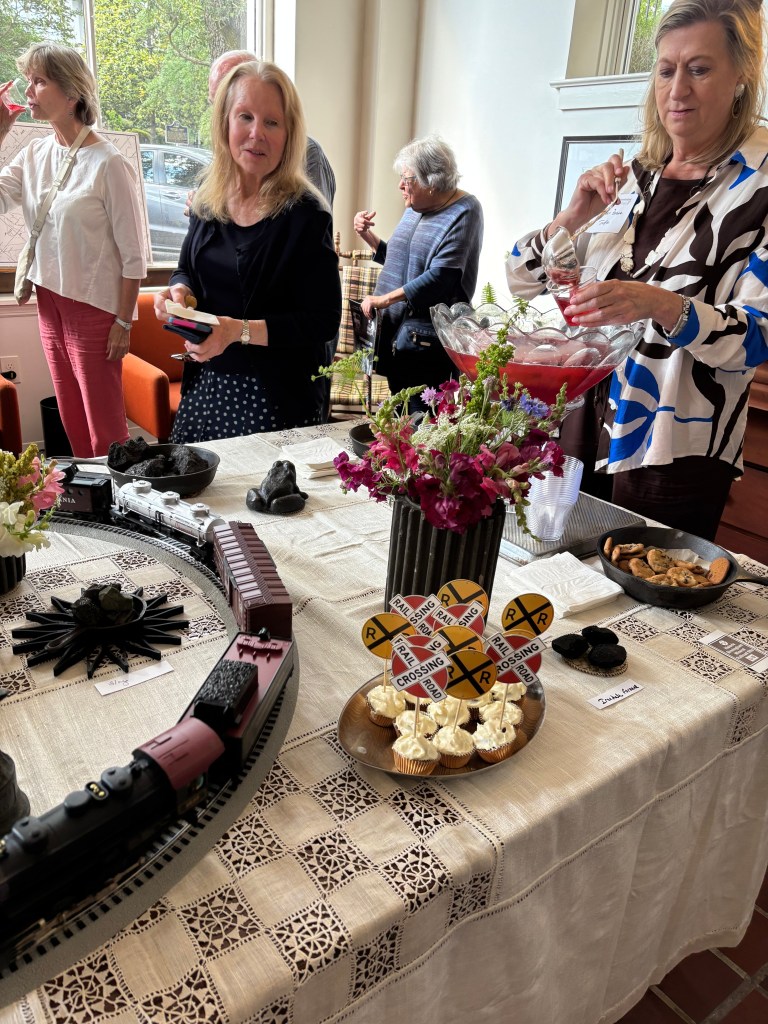 A table set with snacks and drinks, featuring a model train track, floral arrangements, and decorated cupcakes with railway signs. Guests are mingling in the background.